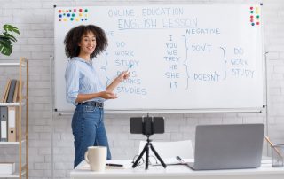 Online English lessons at home. Smiling african american teacher points to white board with rules and records video for students in living room interior with laptop and smartphone